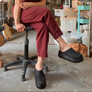 Person wearing maroon pants and black clogs sitting on a stool in a workshop.